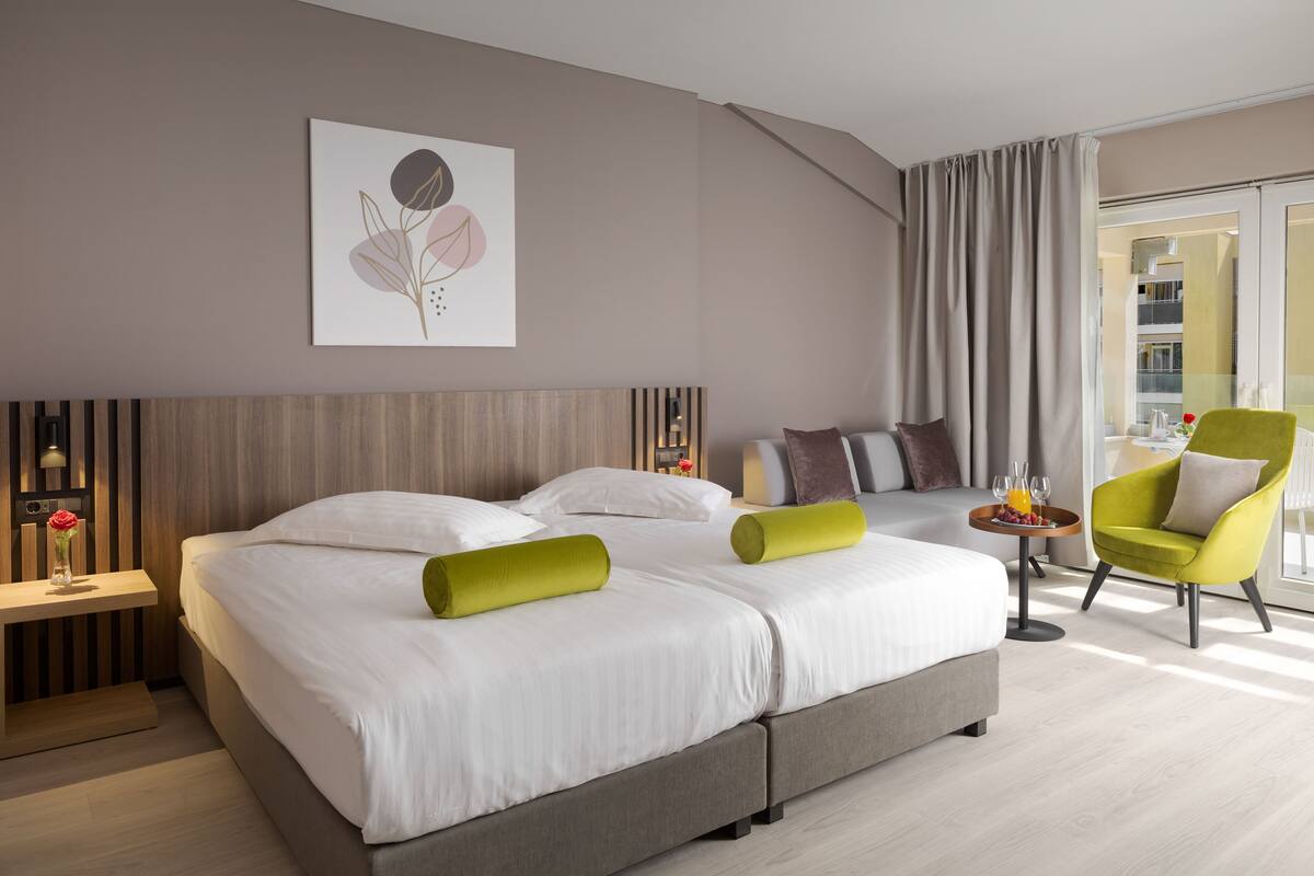 Ein modernes Hotelzimmer mit einem großen Bett, einem kleinen Beistelltisch, einem leuchtend gelben Sessel und einem großen Fenster mit Vorhängen.
