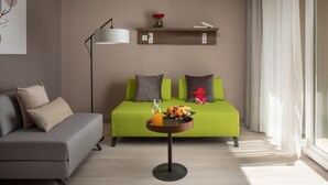 Een moderne woonkamer met een groene bank, een grijze bank, een bijzettafeltje met een fruitschaal en een glas sinaasappelsap, een vloerlamp en een plank met boeken en bloemen.