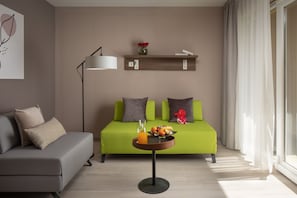 Un salon moderne avec un canapé vert, un canapé gris, une petite table sur laquelle se trouve un panier de fruits et un verre de jus d’orange, une lampe sur pied et une étagère avec des livres et des fleurs.