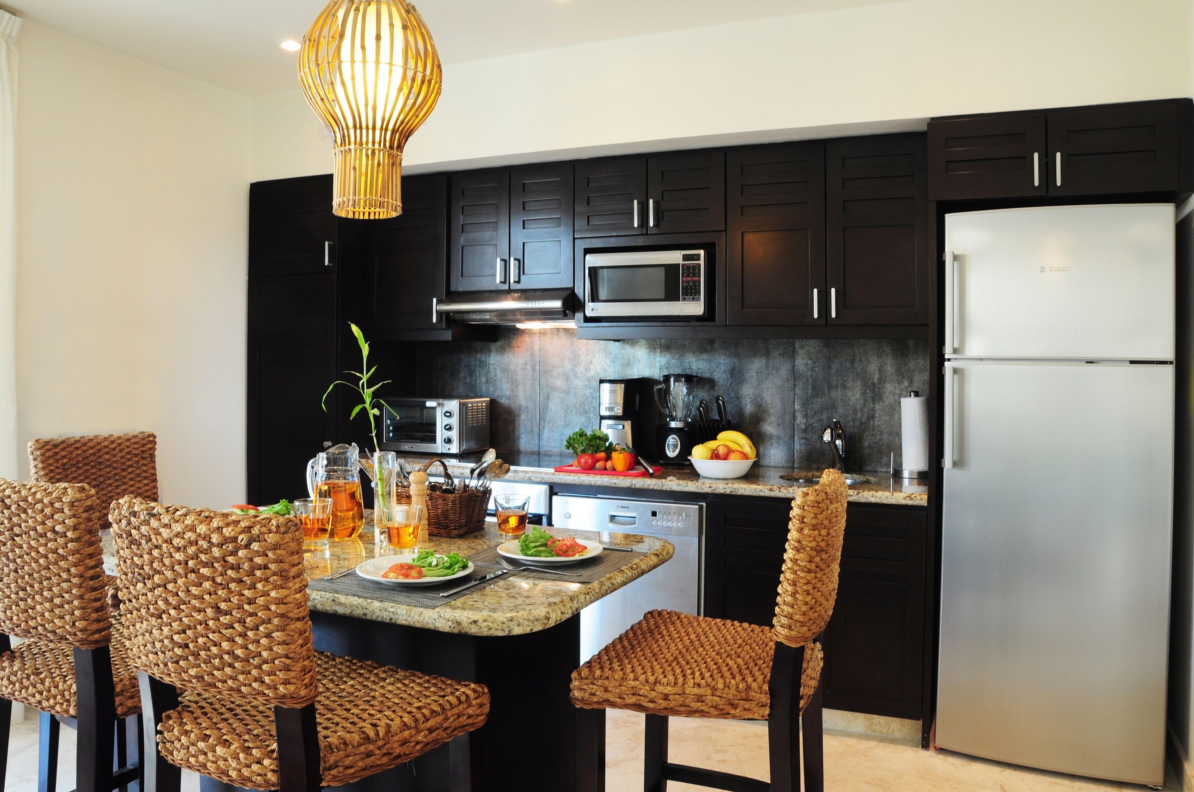 A modern kitchen with dark cabinets, a stainless steel refrigerator, a microwave, and a coffee maker. There is a dining table with wicker chairs and a hanging light fixture.