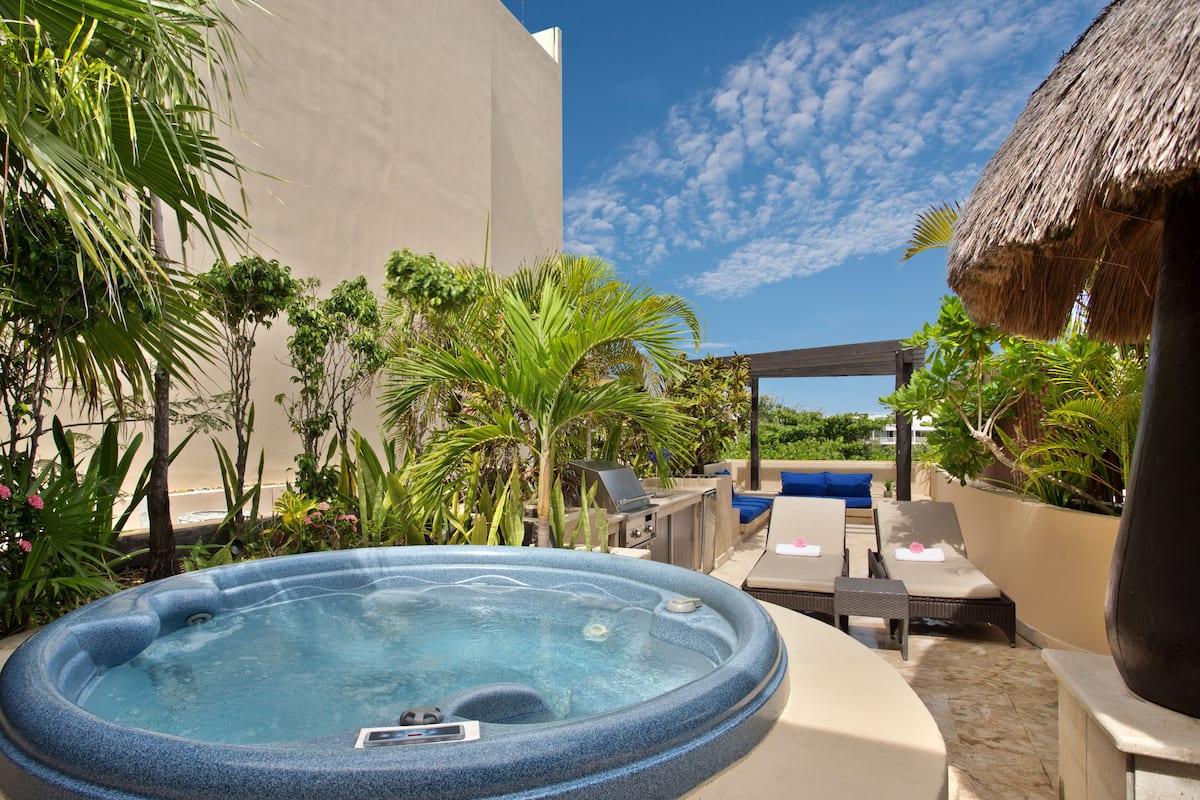 A hot tub surrounded by lounge chairs and a barbecue area, with a thatched roof structure and tropical plants in the background.