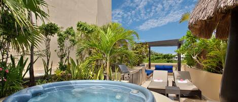 A hot tub surrounded by lounge chairs and a barbecue area, with a thatched roof structure and tropical plants in the background.