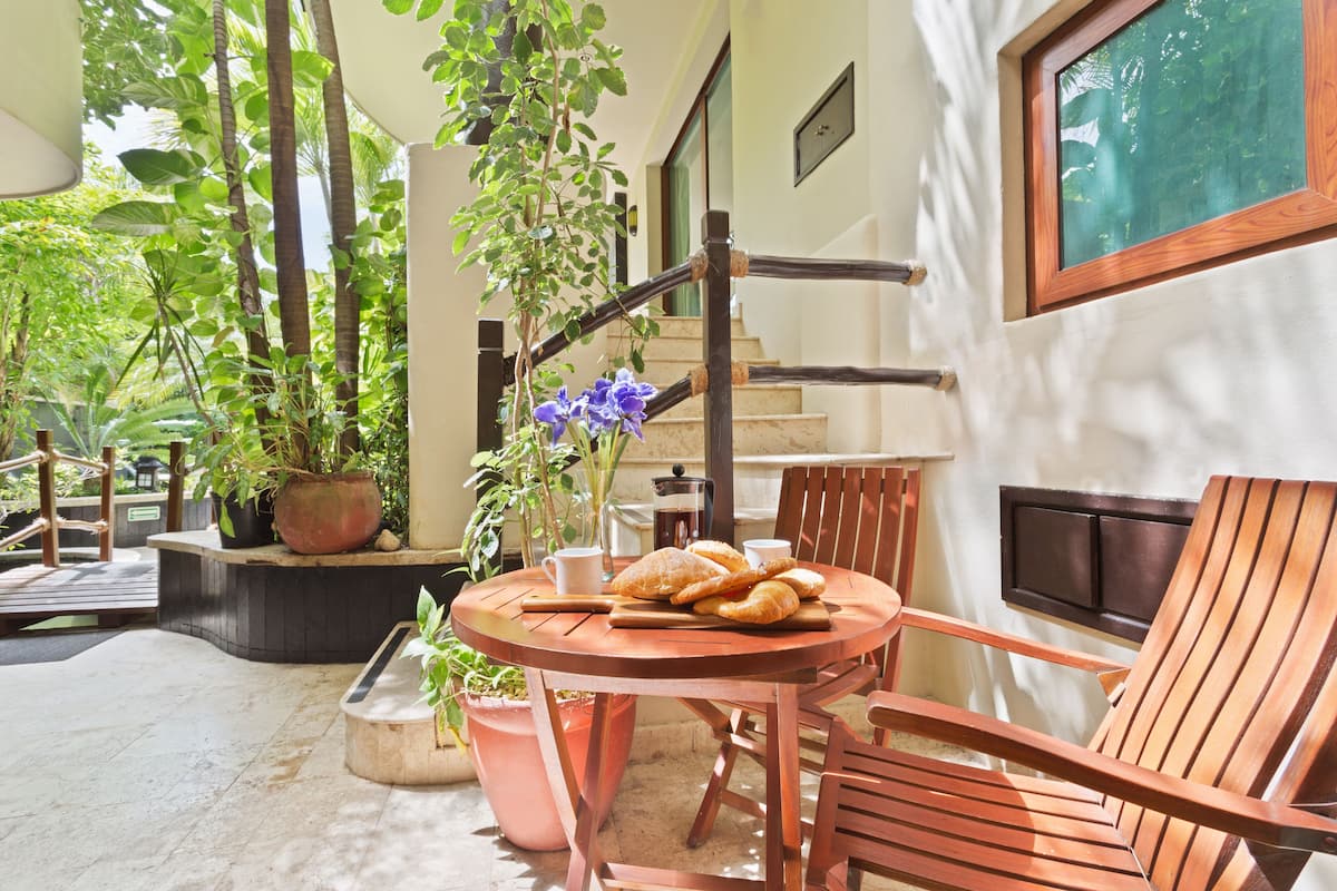 A wooden table with bread and a coffee pot, surrounded by chairs, in a sunny outdoor area with potted plants and a staircase.