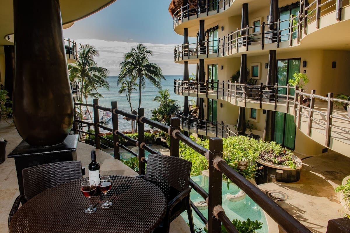 A balcony with a table, two wine glasses, and a bottle of wine. The balcony overlooks a pool and a building with multiple balconies.