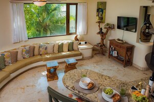 Una sala de estar con un sofá curvo, una mesa de comedor con desayuno servido, un televisor y un ventanal grande con cortinas.