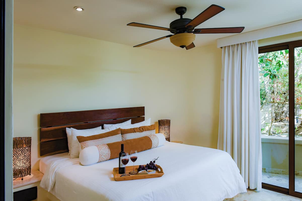 A hotel room with a bed, a ceiling fan, a nightstand, a tray with wine glasses and grapes, and a view of greenery outside.