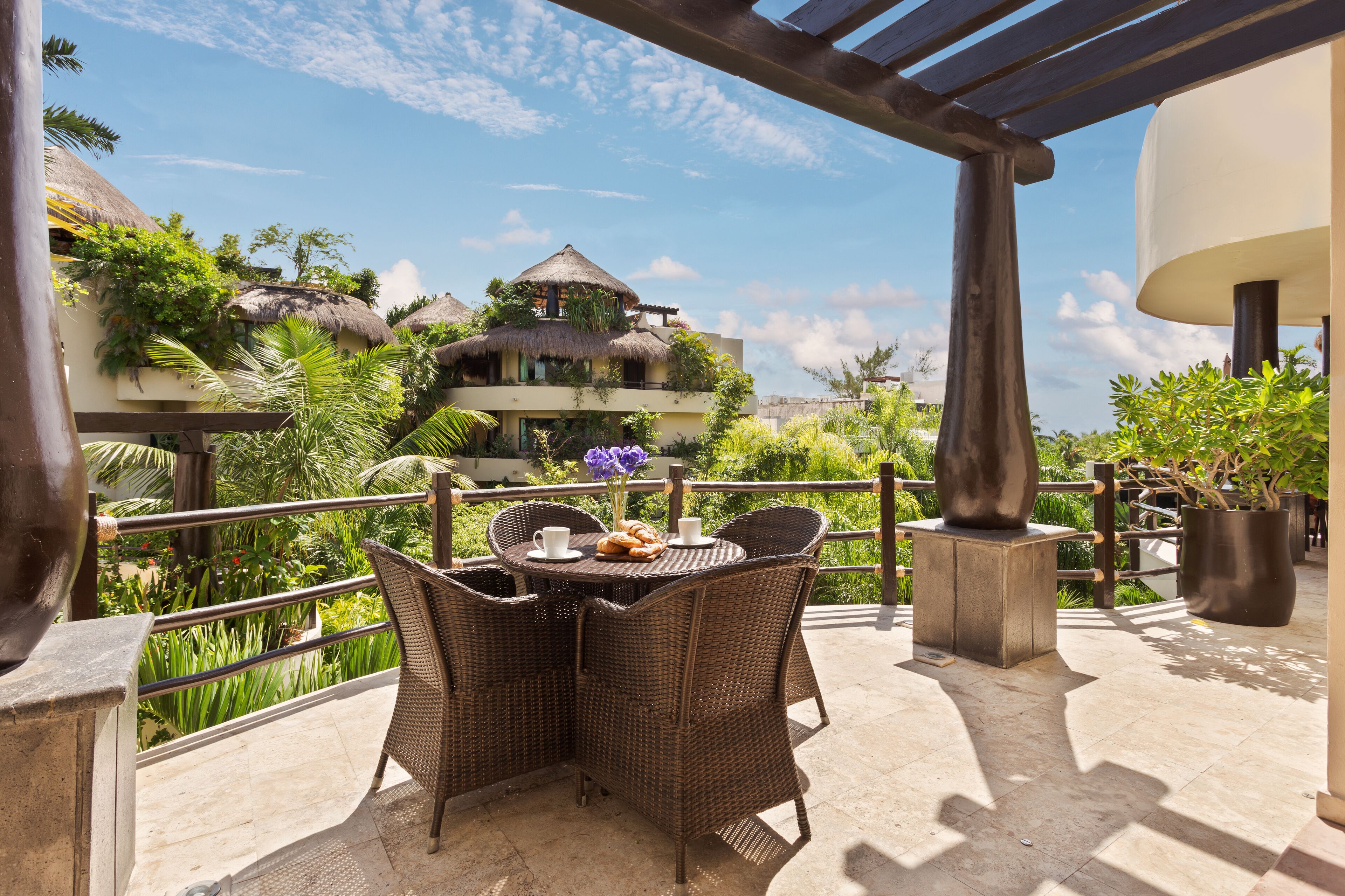 A terrace with wicker furniture, a table with a cup and pastries, and a view of a building with a thatched roof.