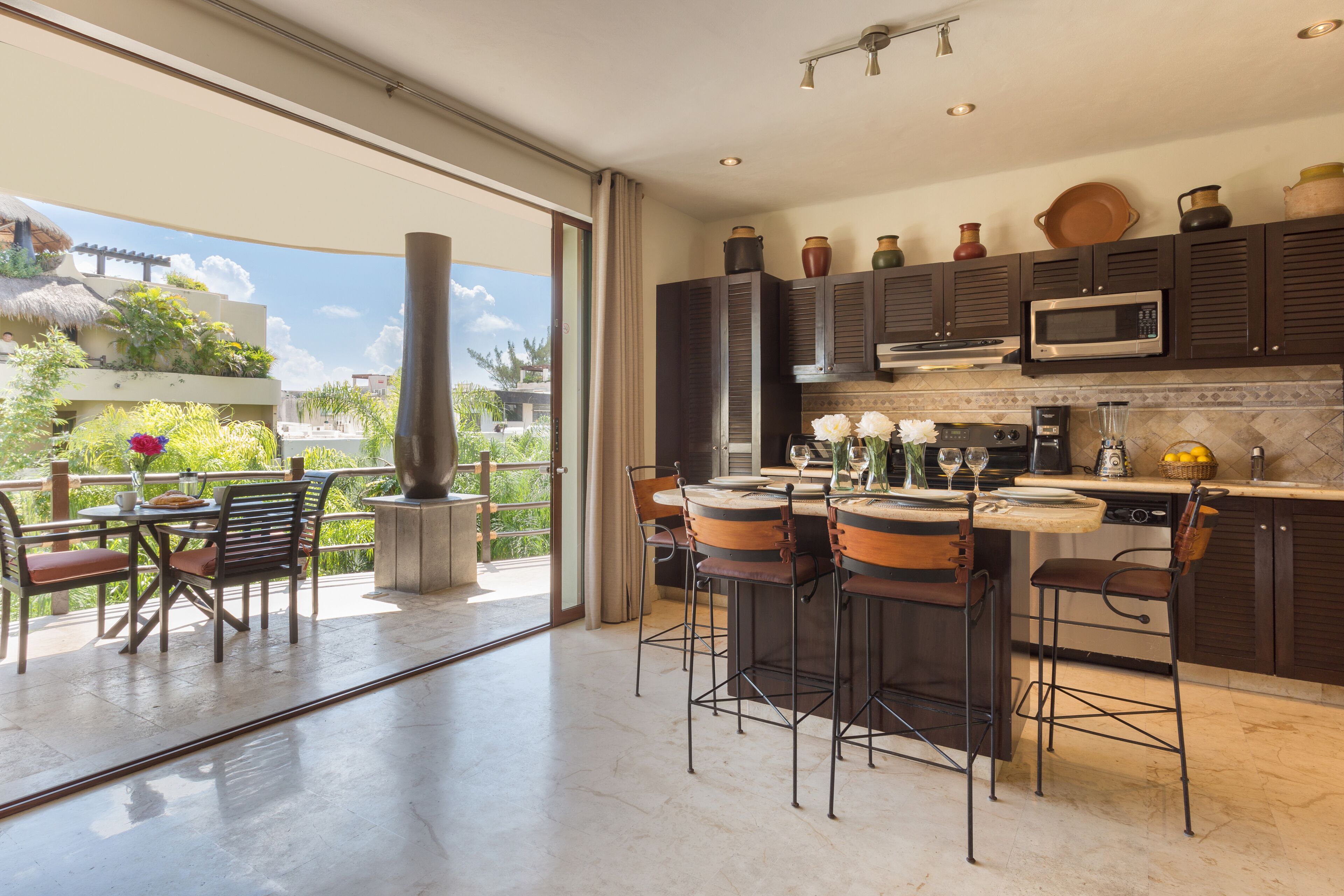 A modern kitchen with dark cabinetry, a central island with bar stools, a large window with a view of greenery, and a dining area with a table and chairs.