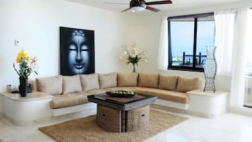 A modern living room with a beige sectional sofa, a coffee table, a large Buddha artwork, a vase with flowers, and a view through a large window.