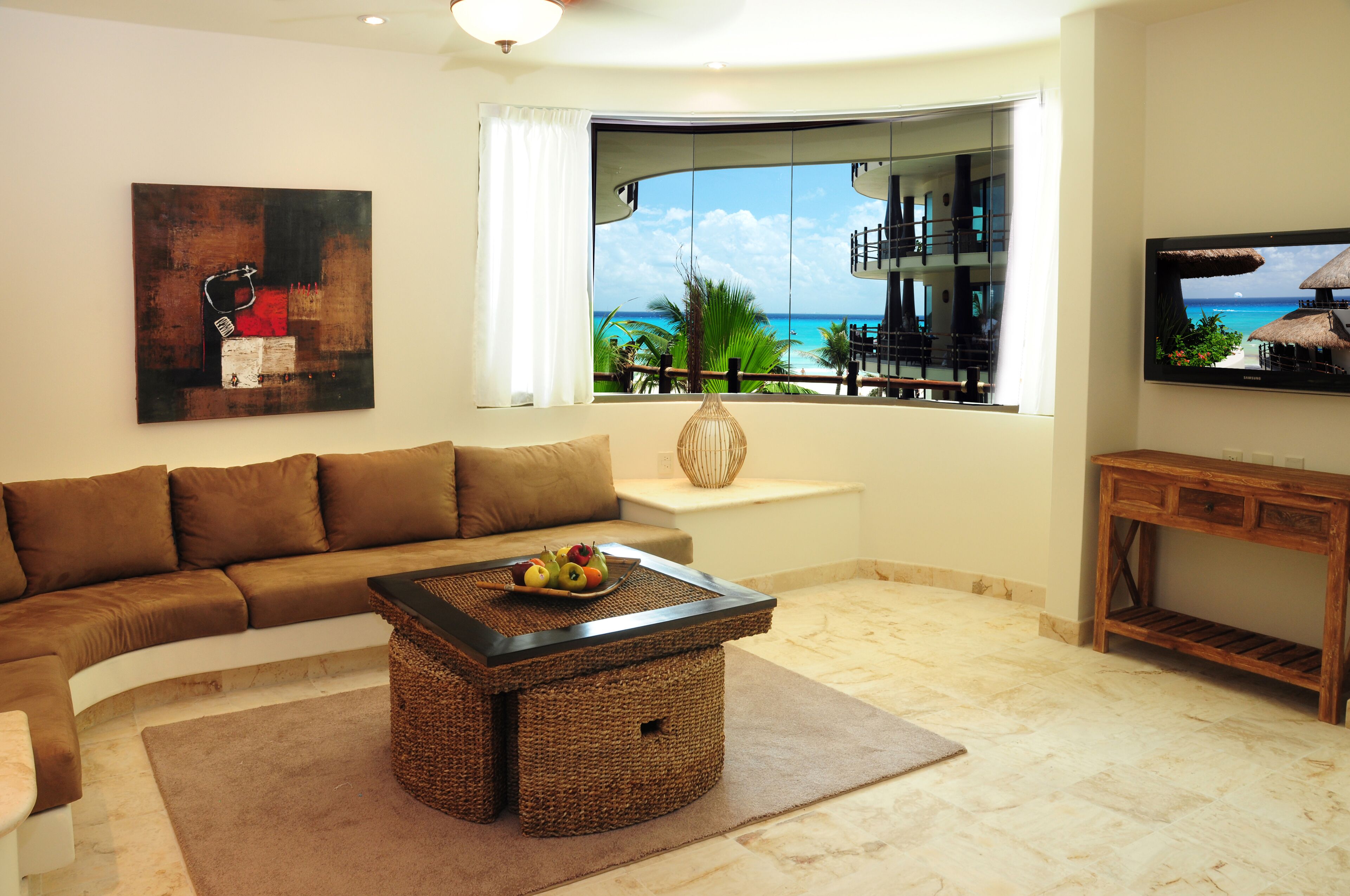A living room with a large window, a brown sofa, a coffee table with fruit, and a TV on the wall.