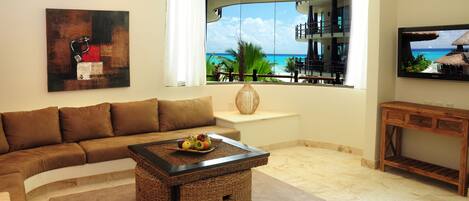 A living room with a large window, a brown sofa, a coffee table with fruit, and a TV on the wall.