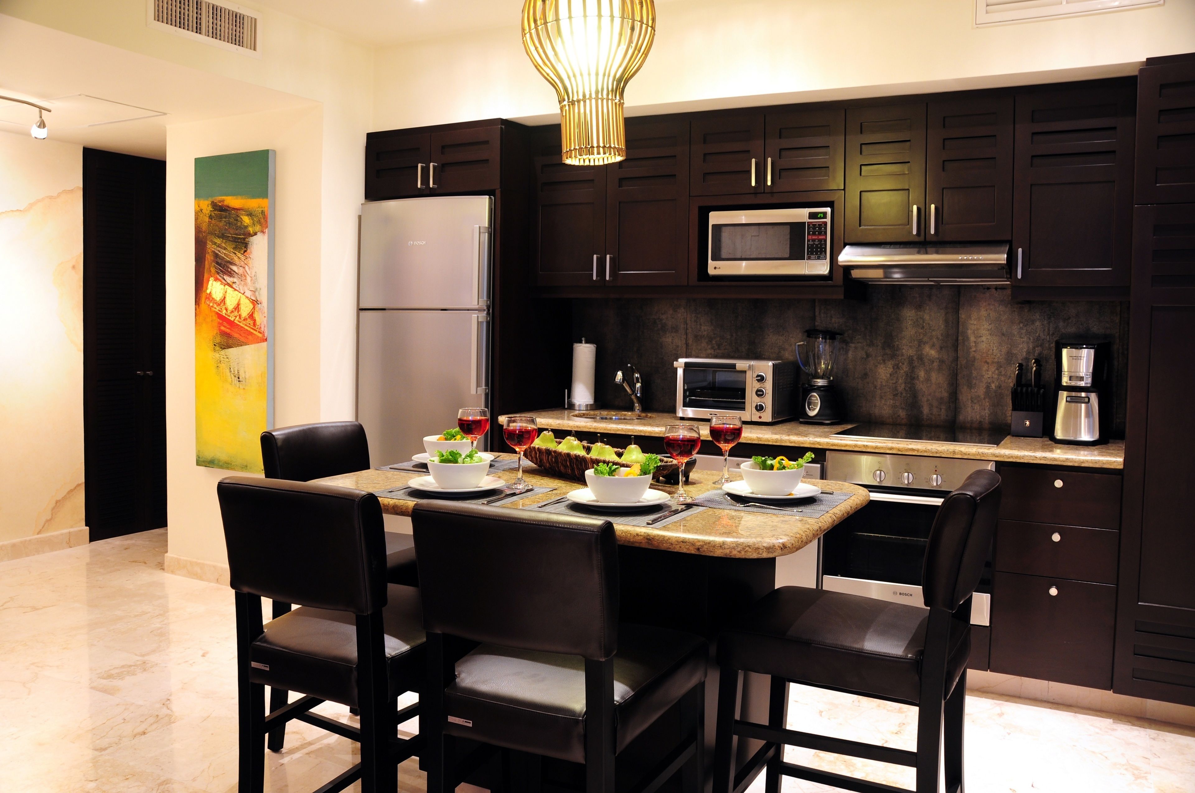 A modern kitchen with dark cabinets, a stainless steel refrigerator, a microwave, and a coffee maker. There is a dining table with four chairs and a painting on the wall.