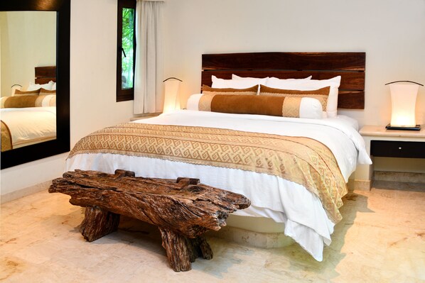 Una cama bien hecha con cabecera de madera, una mesita de noche con lámpara y un banco rústico de madera en una habitación luminosa.
