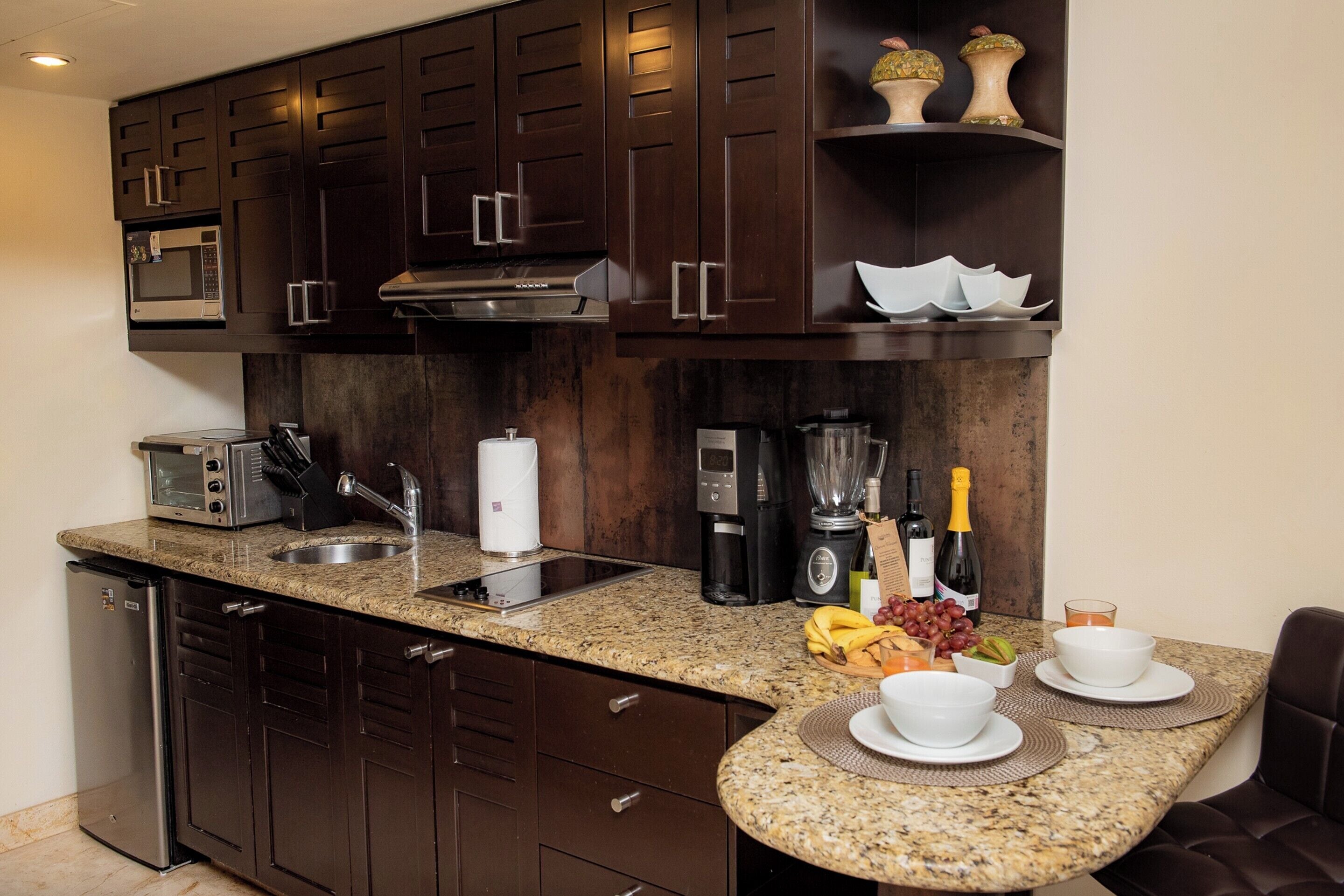 A compact kitchen with dark wood cabinets, a granite countertop, a built-in microwave, a coffee maker, and a wine bottle.