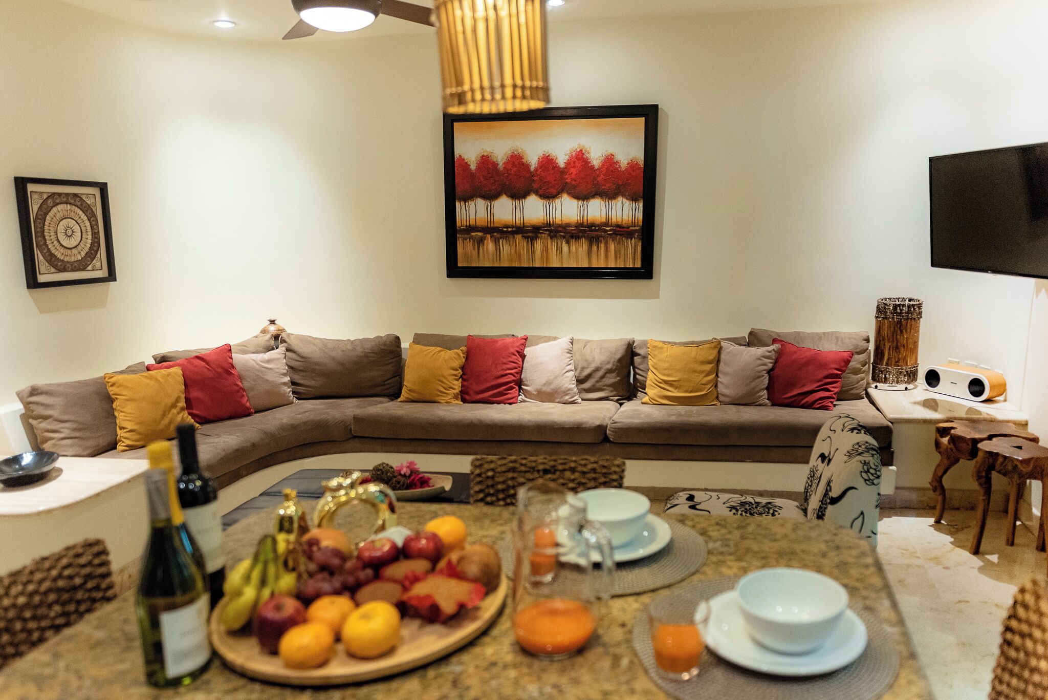 A living room with a sofa, a dining table with fruit and drinks, a painting, and a television.