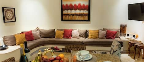 A living room with a sofa, a dining table with fruit and drinks, a painting, and a television.