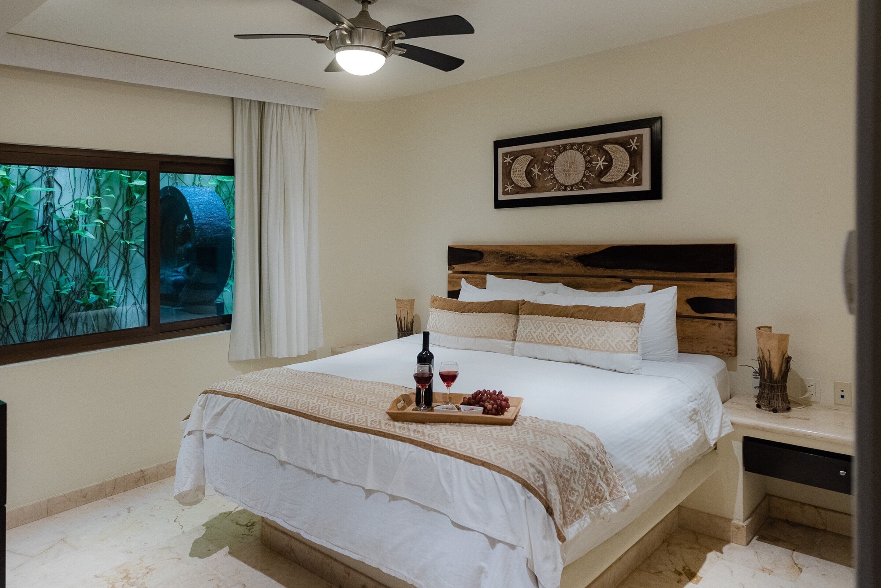 A bedroom with a large bed, a ceiling fan, a window with a view of greenery, and a wall-mounted artwork.