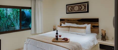 A bedroom with a large bed, a ceiling fan, a window with a view of greenery, and a wall-mounted artwork.