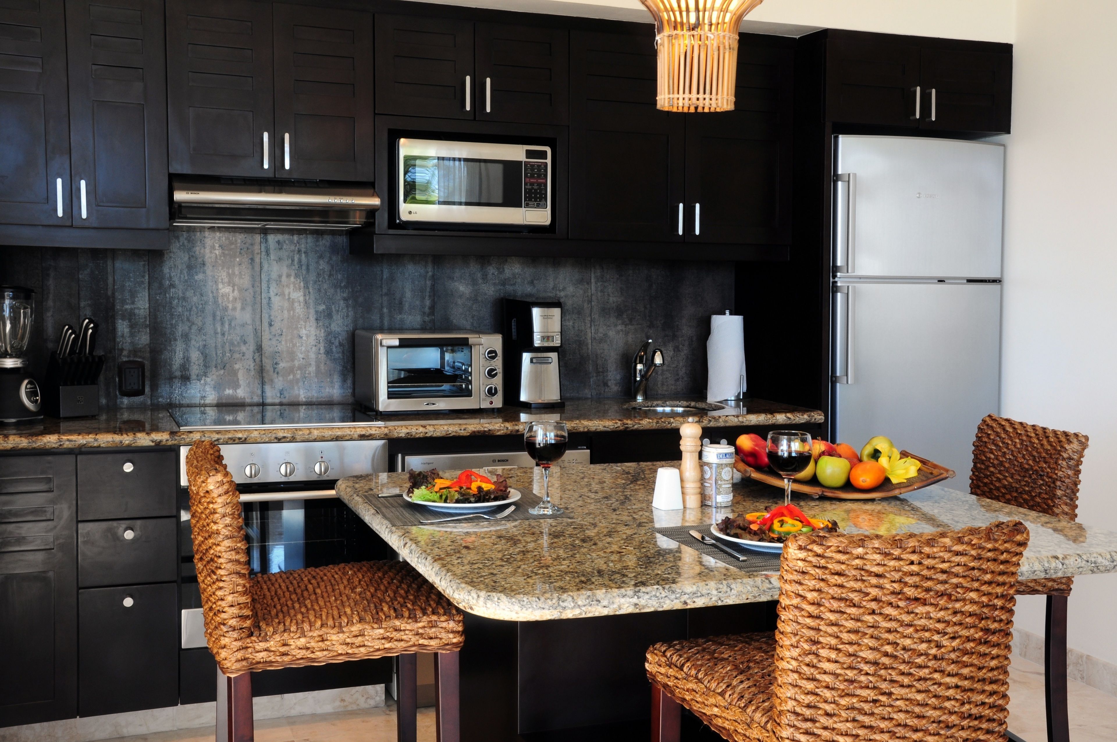 A modern kitchen with dark cabinets, stainless steel appliances, a granite countertop, and wicker chairs.