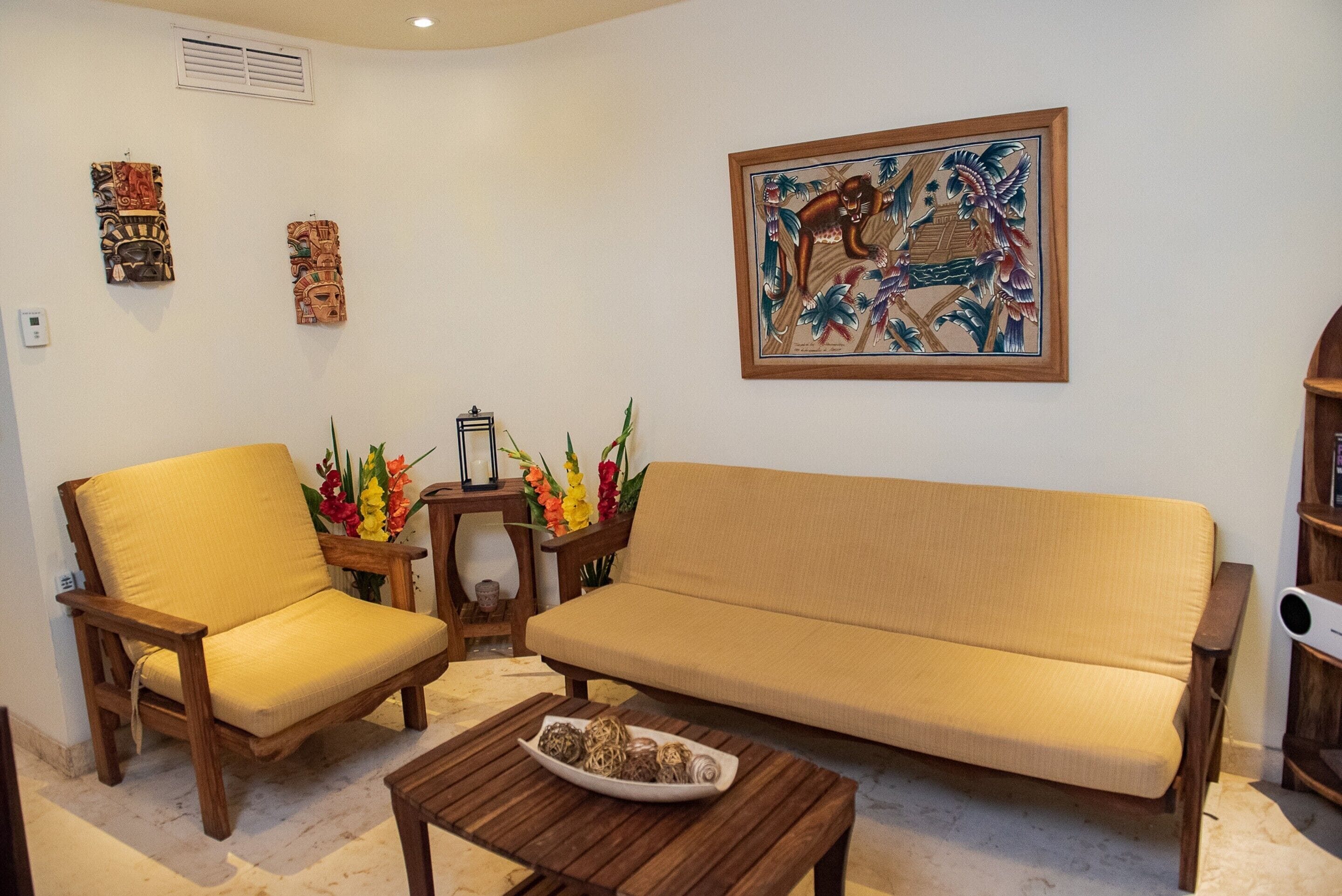 Una sala de estar con un sofá color beige, dos sillas de madera, una mesa de centro con objetos decorativos y obras de arte en la pared.