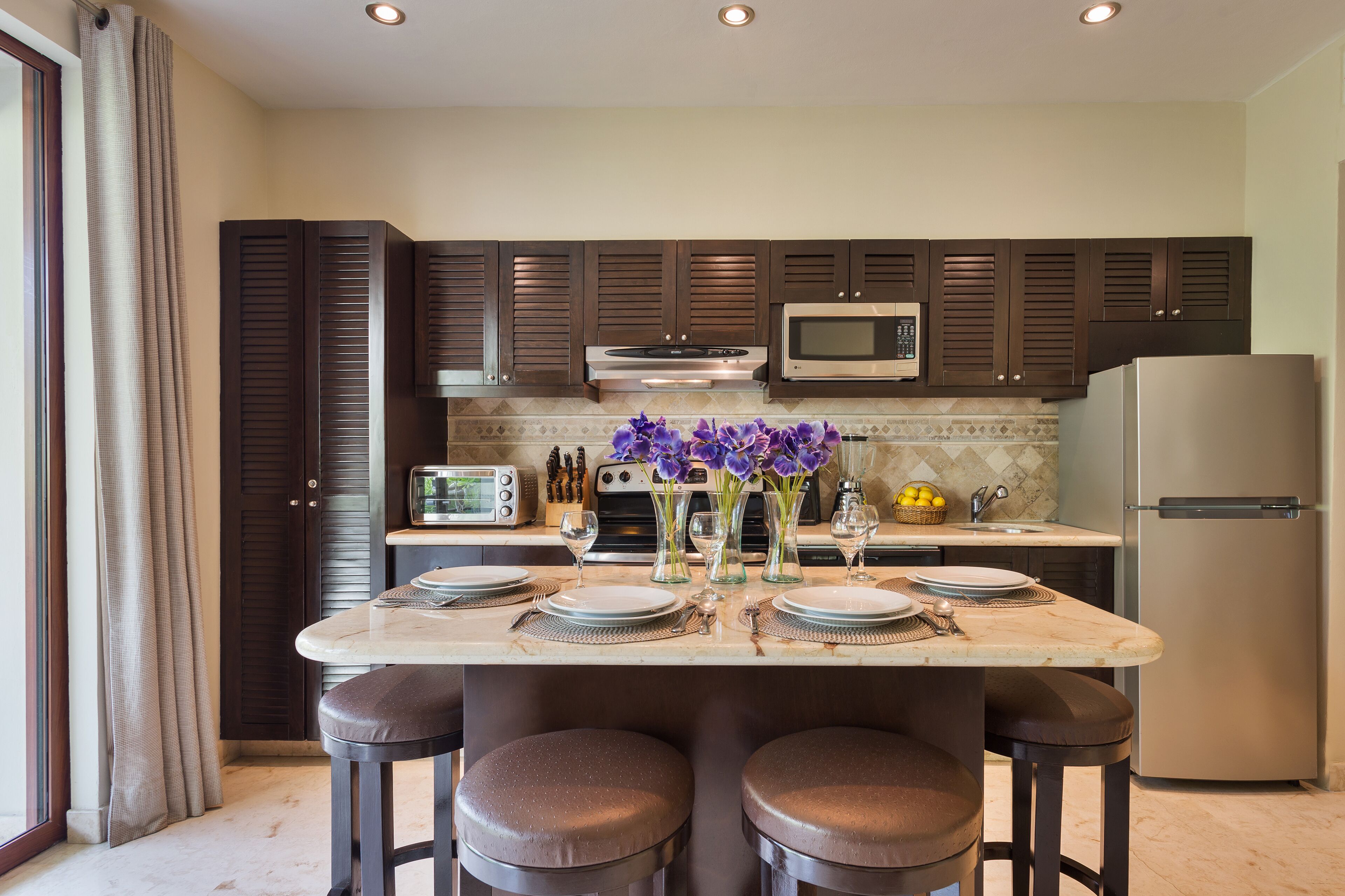 A kitchen with dark cabinets, a microwave, a refrigerator, and a dining table set with plates and glasses.