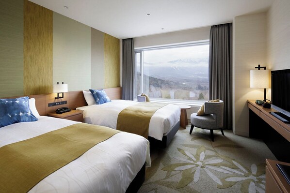 Habitación de hotel con dos camas, un escritorio, una silla, un televisor y un amplio ventanal con vistas a la montaña.