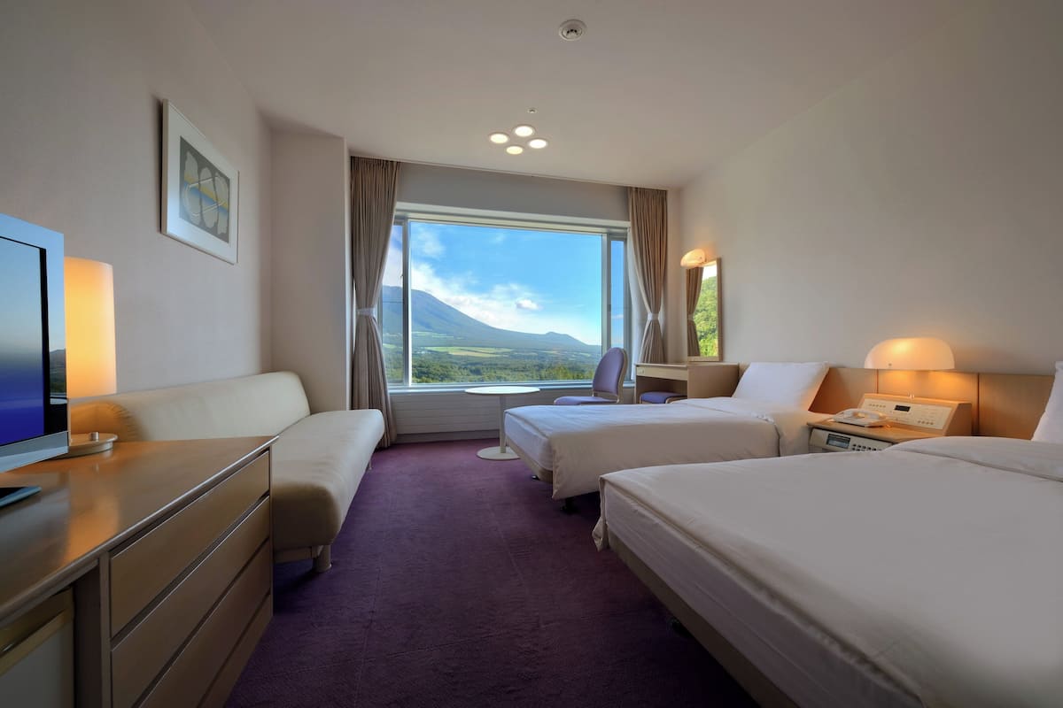 A hotel room with two beds, a desk, a chair, a TV, and a large window with a mountain view.