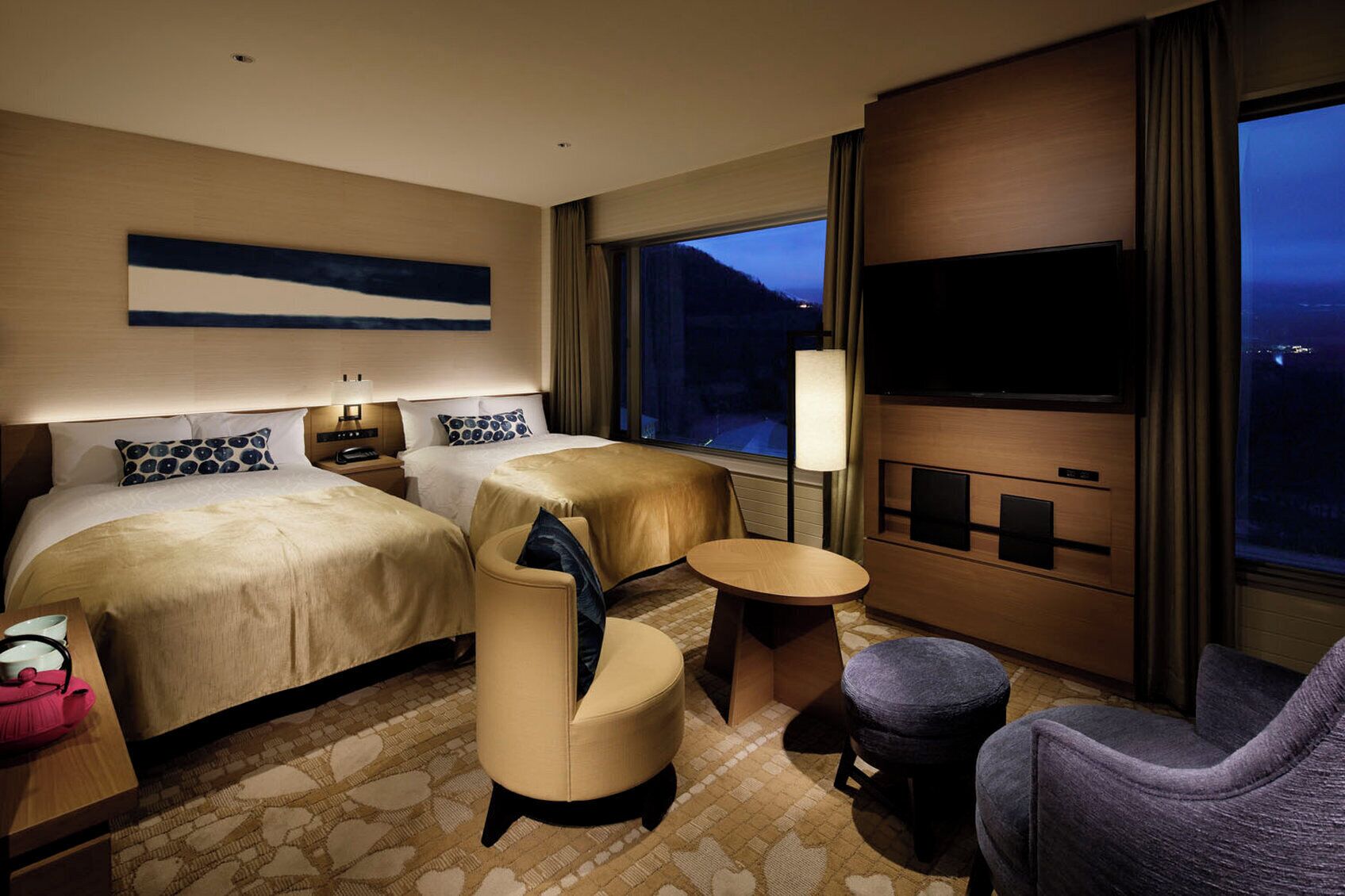 A hotel room with two beds, a TV, a small table, and a view of the city at night.