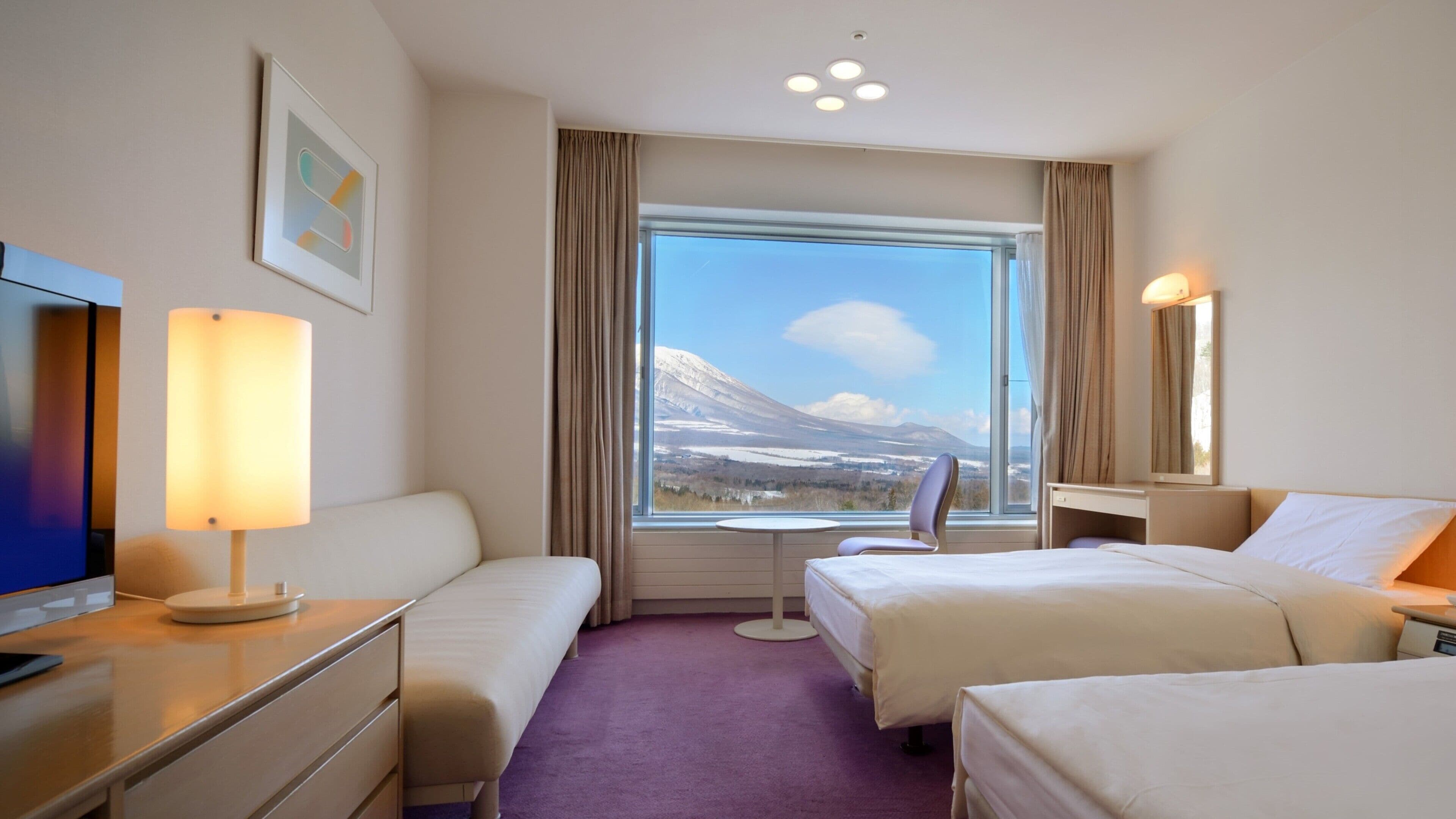 Habitación de hotel con dos camas, un escritorio, una silla, un televisor y un amplio ventanal con vistas a la montaña.