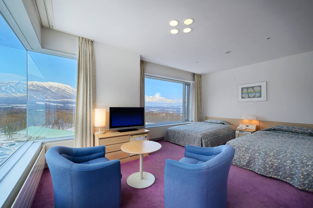 A hotel room with a large window, a bed, a TV, a desk, and two chairs.