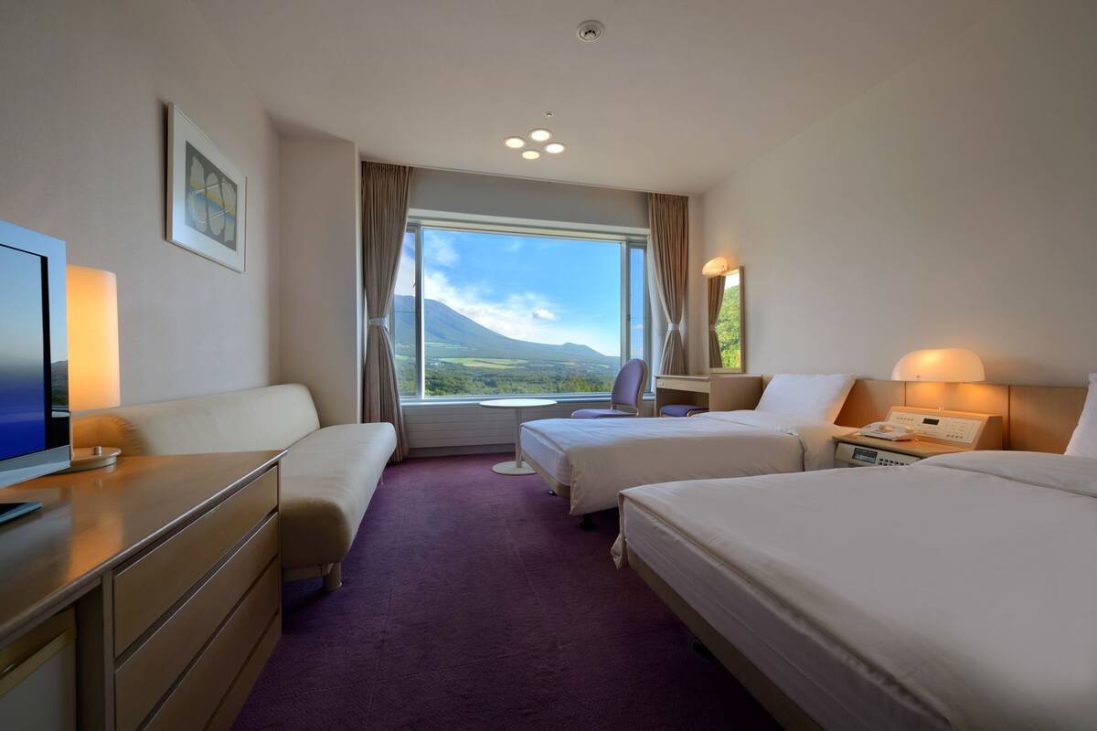 A hotel room with two beds, a desk, a TV, and a large window with a mountain view.