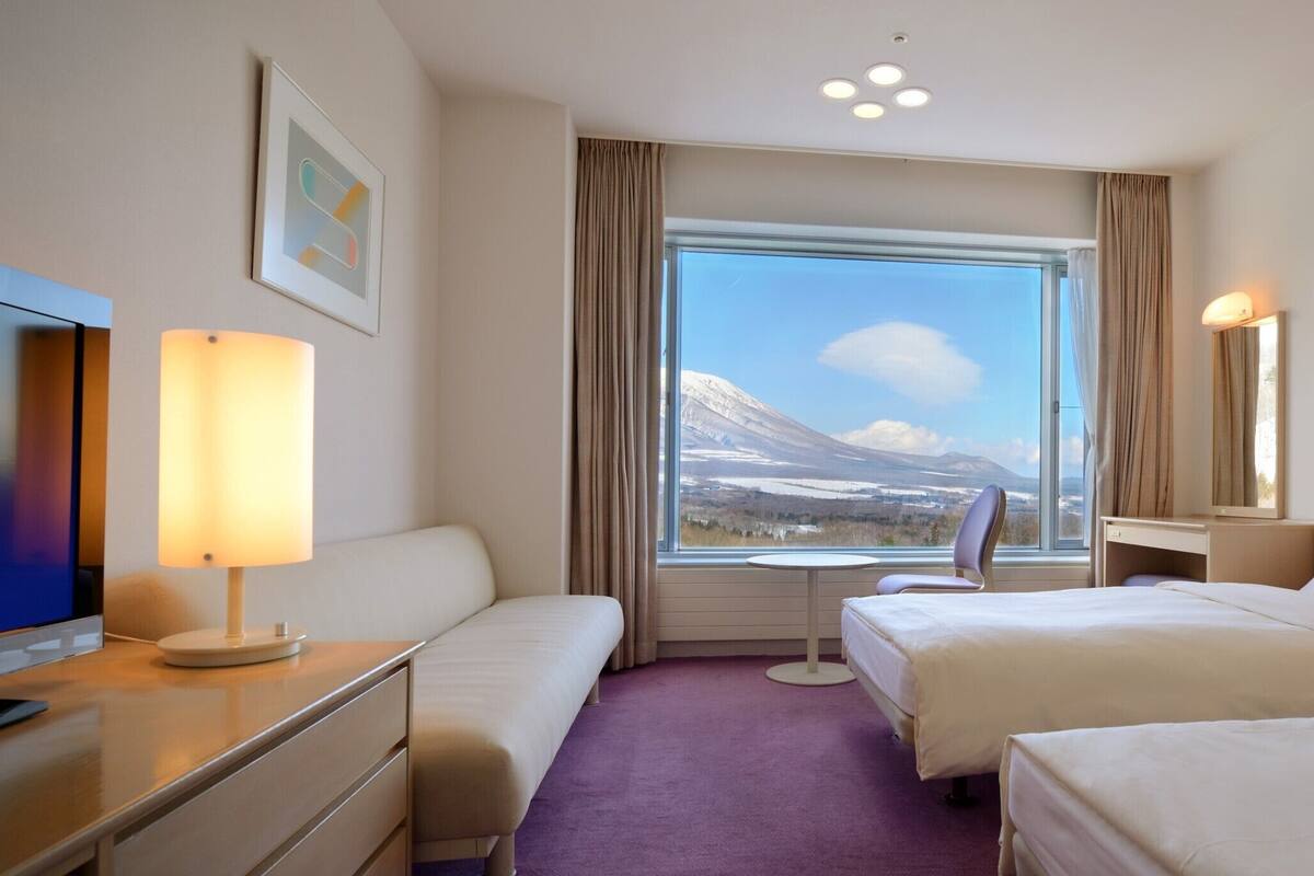 Et hotelværelse med to senge, et skrivebord, en stol, et fjernsyn og et stort vindue med udsigt til et bjerglandskab.