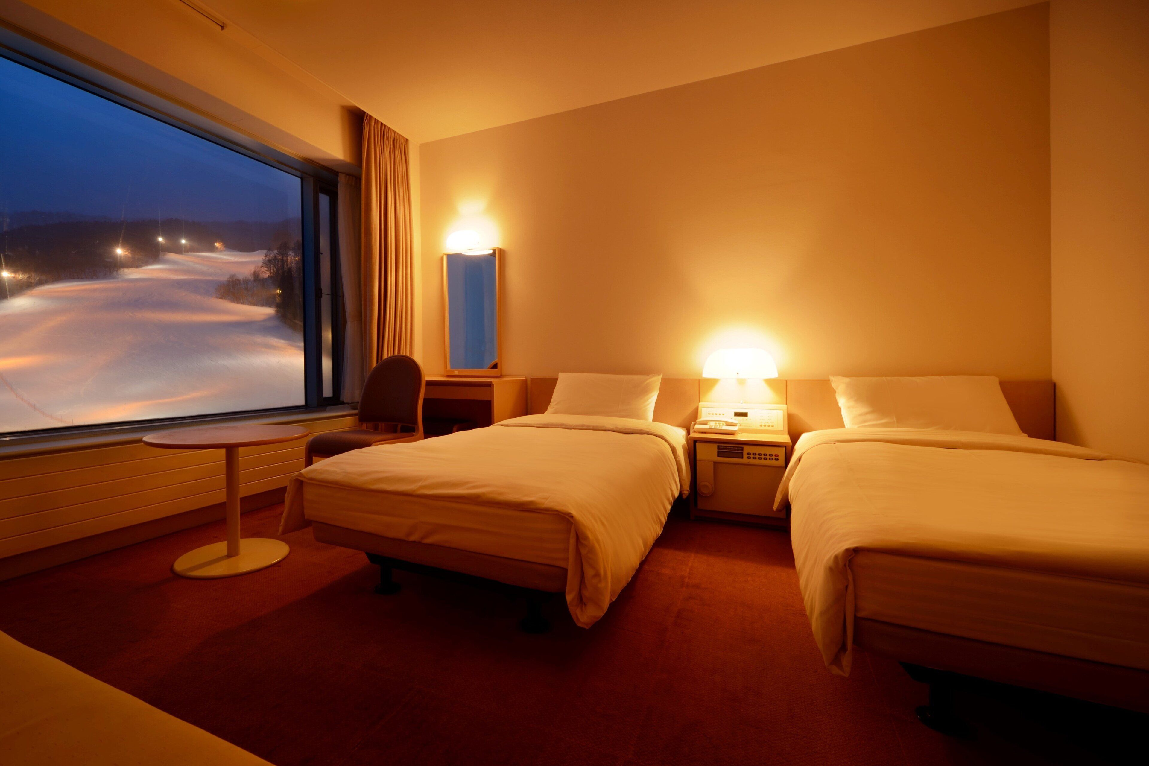 A hotel room with two beds, a small table, a chair, a nightstand with a lamp, and a window with a view of a snowy landscape.