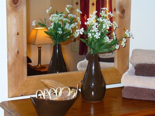 A wooden table with two vases of white flowers, a bowl of white flowers, a lamp, and a mirror reflecting the room.