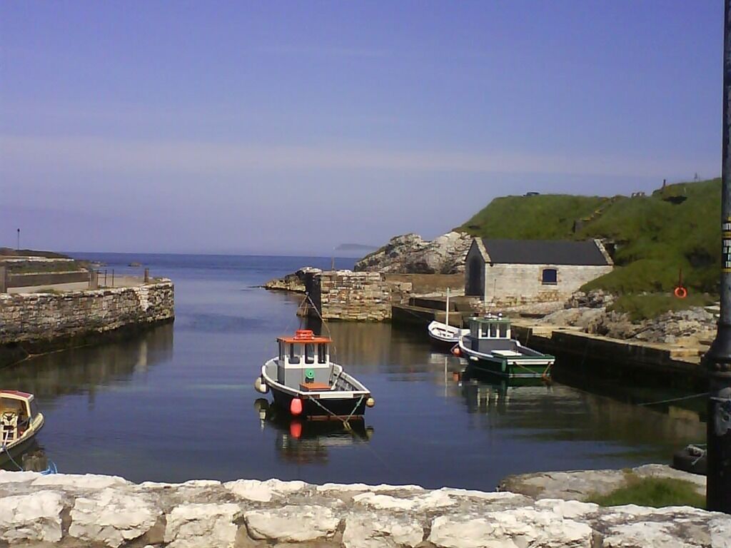 A harbor with boats docked, a stone wall, and a small building with a green roof.