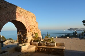 Un antiguo arco de piedra con vistas a la costa, con plantas en macetas y una zona pavimentada.