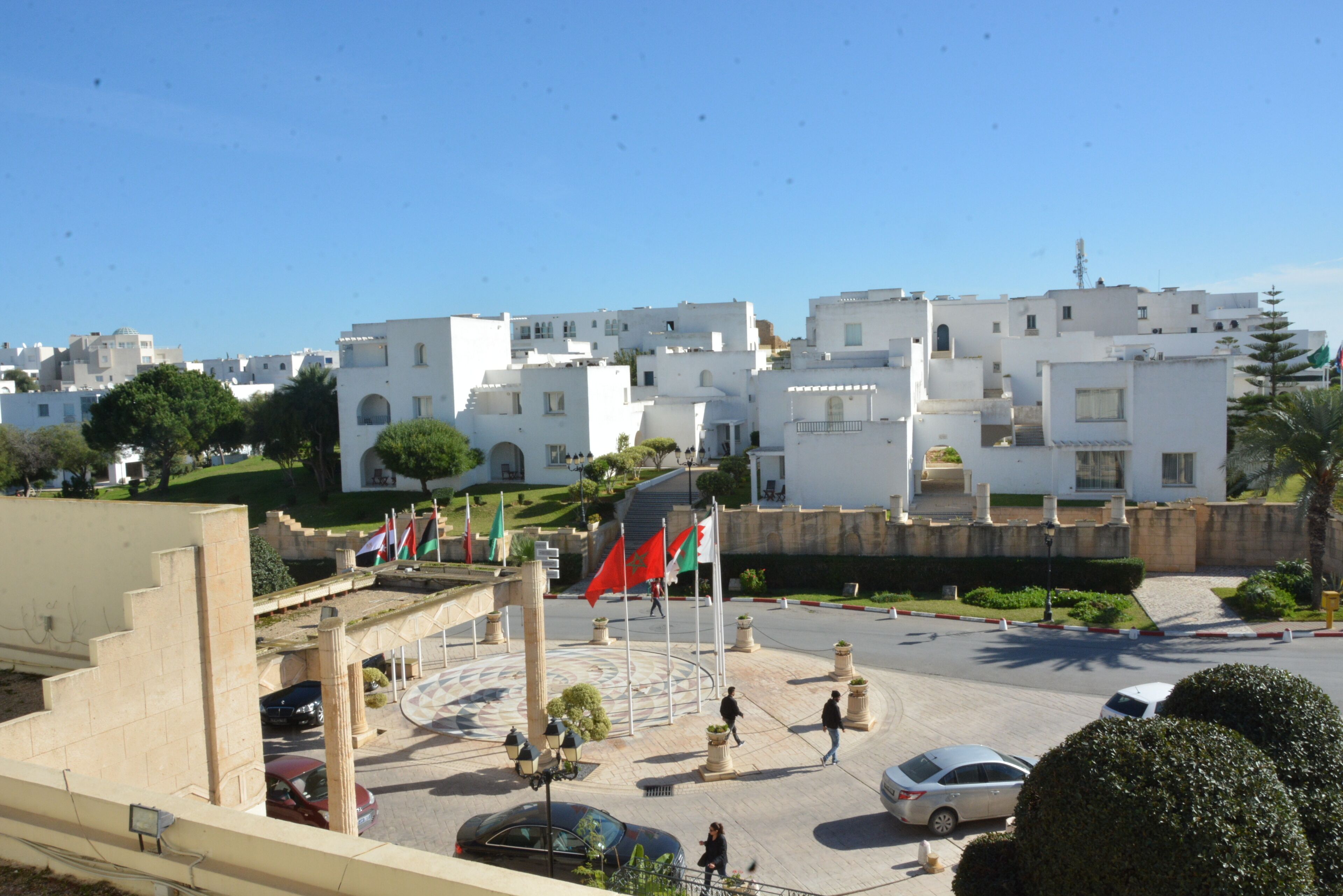 A view of a town with white buildings, a central plaza with flags, and a few cars parked.
