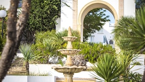 A stone fountain surrounded by lush greenery and a white building with an arched entrance. - Golden Carthage Residence (La Marsa)