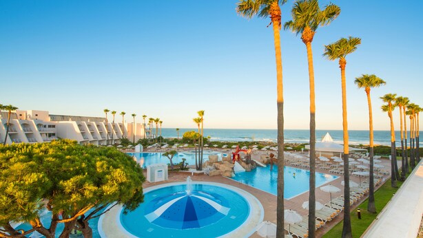 Zwembadgedeelte van een resort met een fontein, ligbedden en palmbomen.