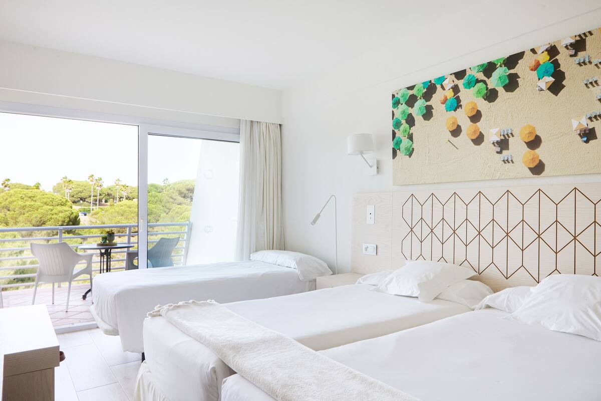 Een hotelkamer met twee bedden, een balkon met een tafel en stoelen, en uitzicht op groen.