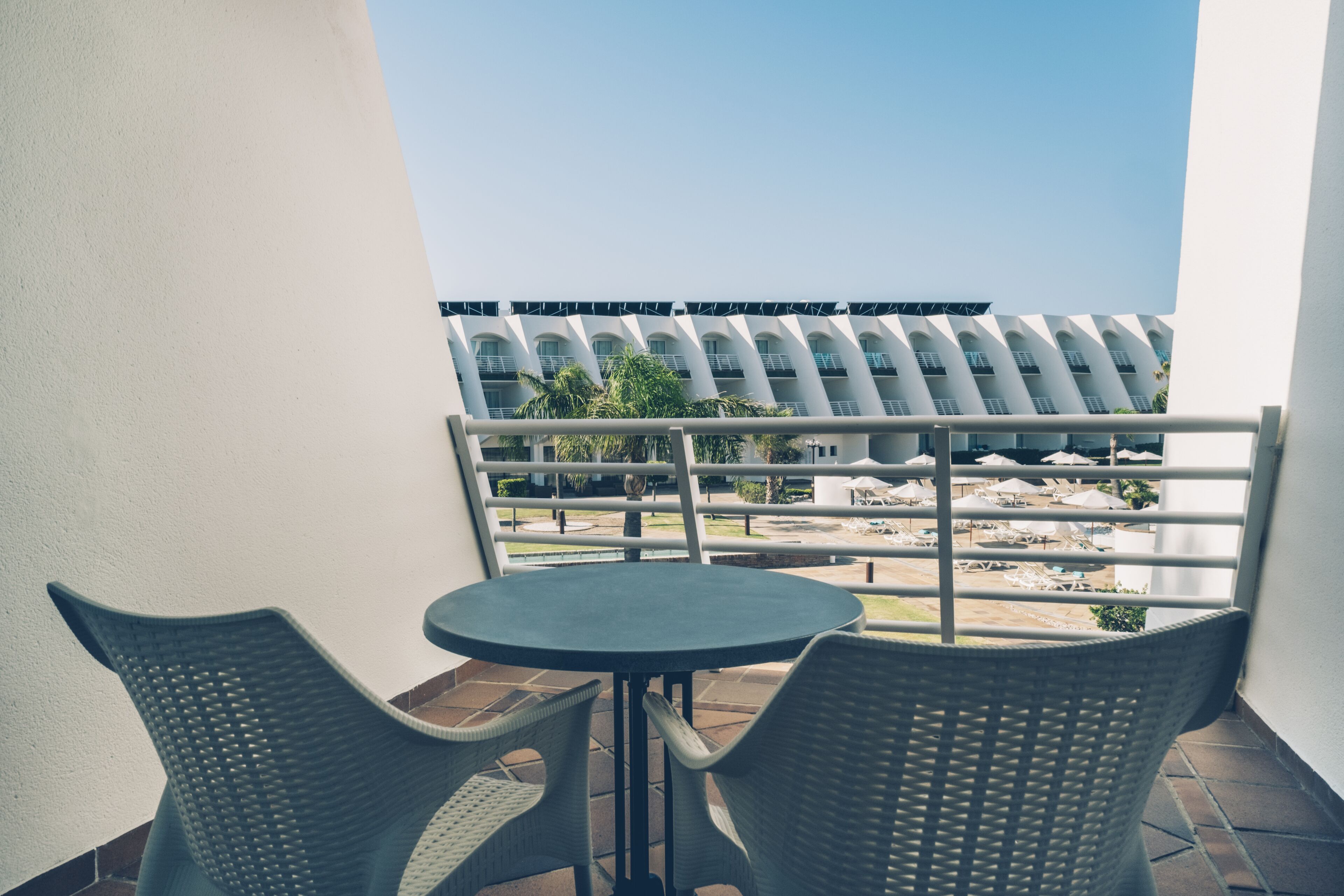 A balcony with two wicker chairs and a small table, overlooking a building with a modern design and a pool area.