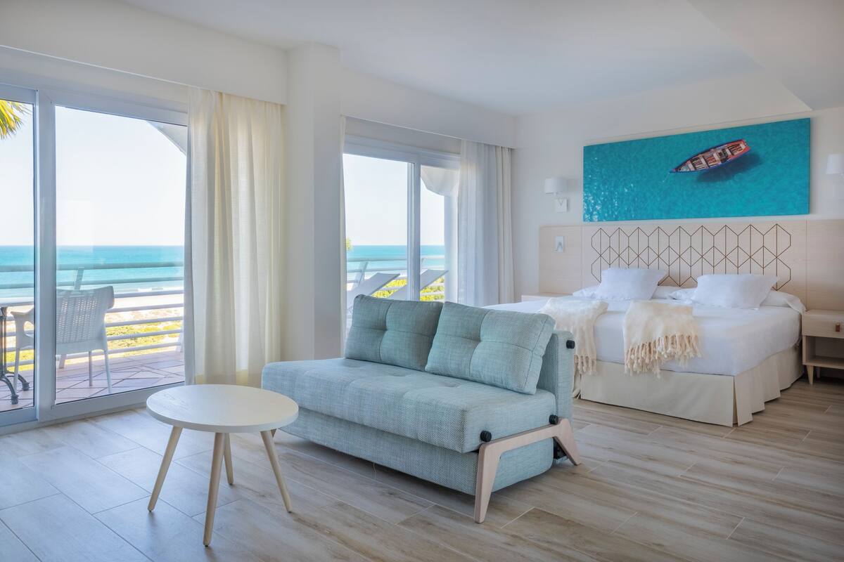 Een hotelkamer met een groot raam en uitzicht op zee, een bed met wit beddengoed, een grijze bank en een kleine ronde tafel.