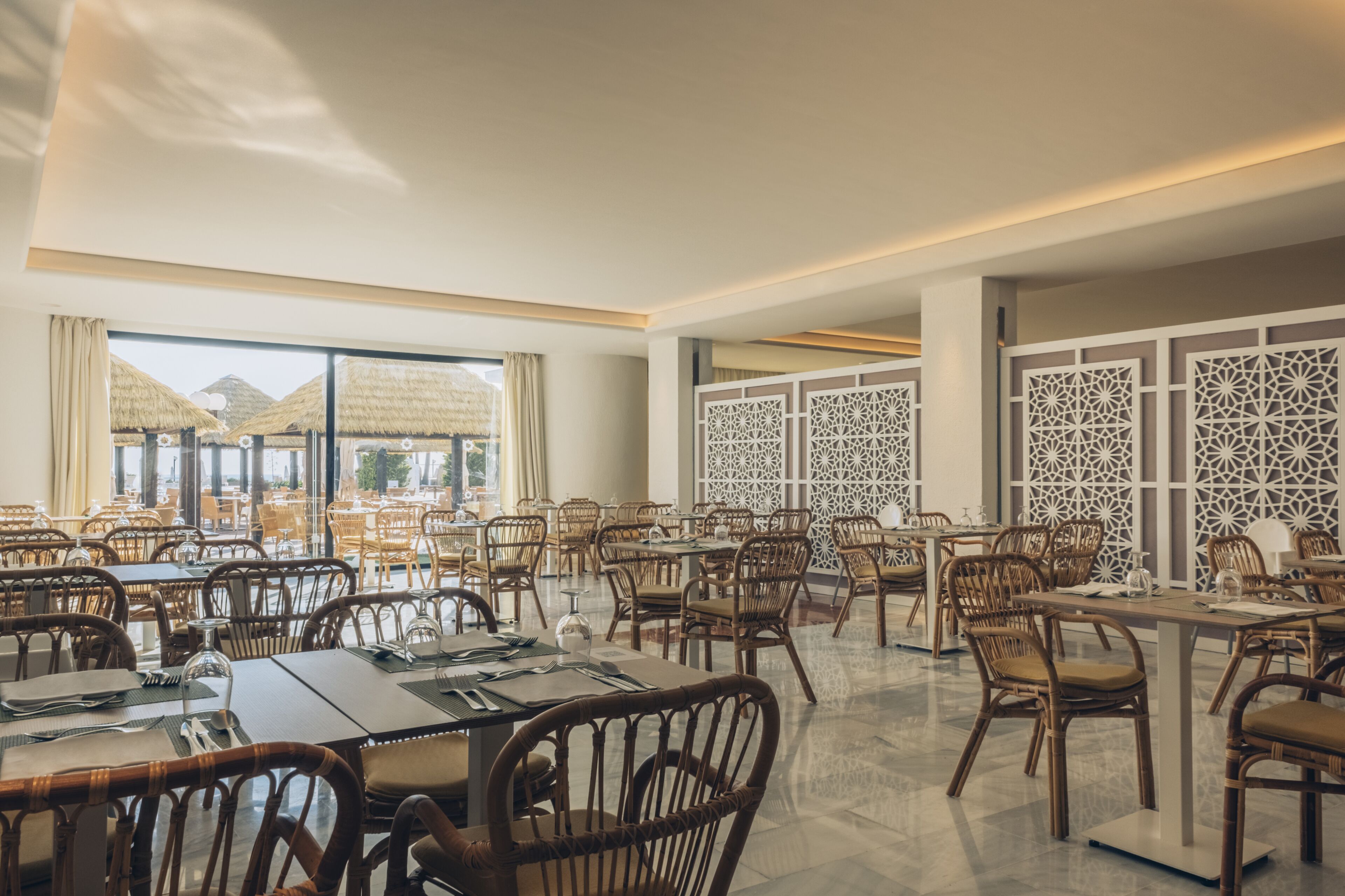 Un comedor amplio con varias mesas y sillas, ventanas grandes que ofrecen vistas de edificaciones con techo de paja en el exterior, y paneles blancos decorativos en las paredes.