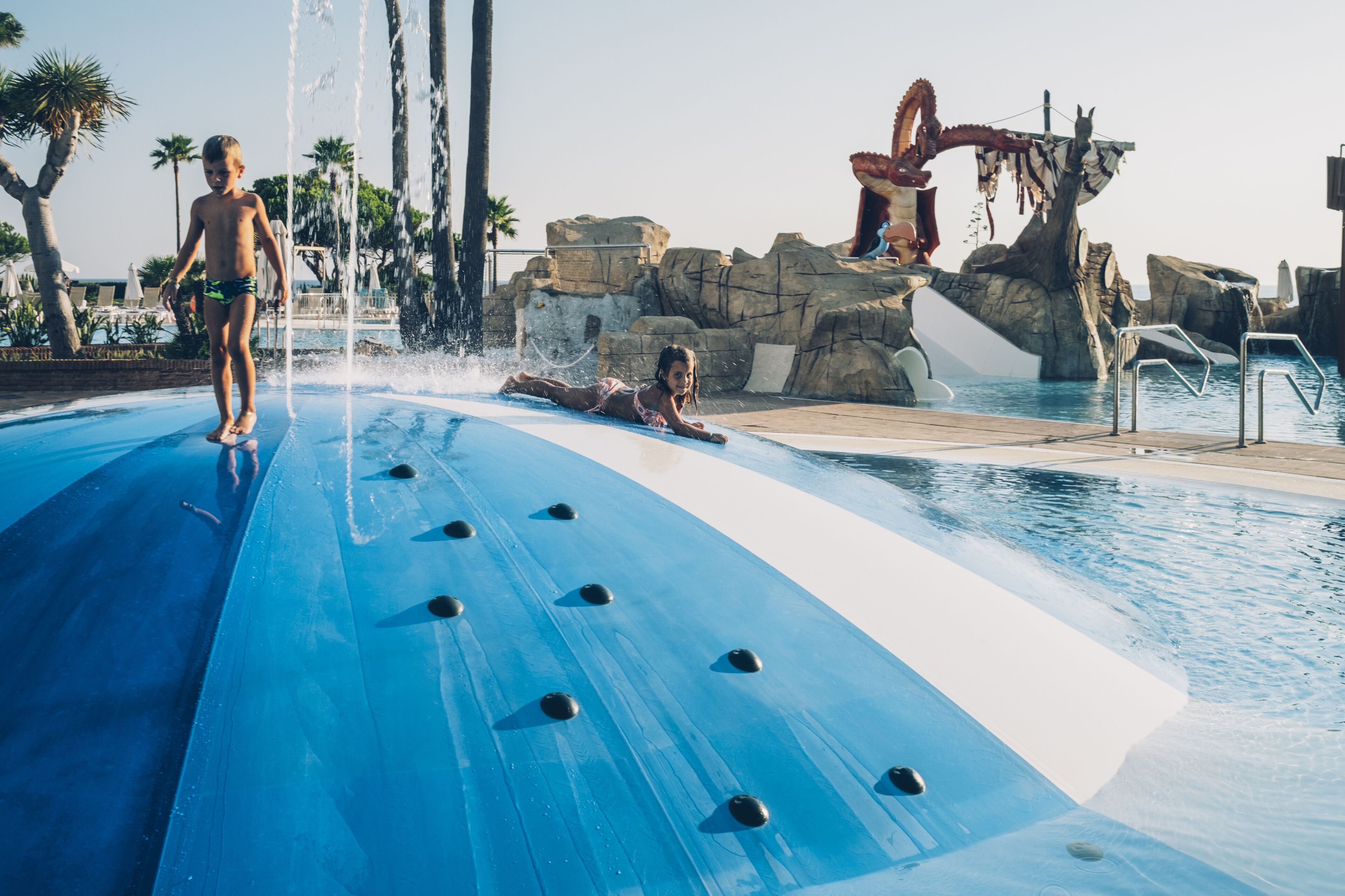 Un tobogán con superficie azul y remaches negros, una estructura rocosa con un brazo mecánico rojo y un área de piscina con palmeras.