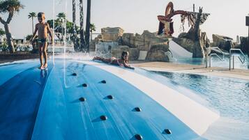 Un tobogán con superficie azul y remaches negros, una estructura rocosa con un brazo mecánico rojo y un área de piscina con palmeras.