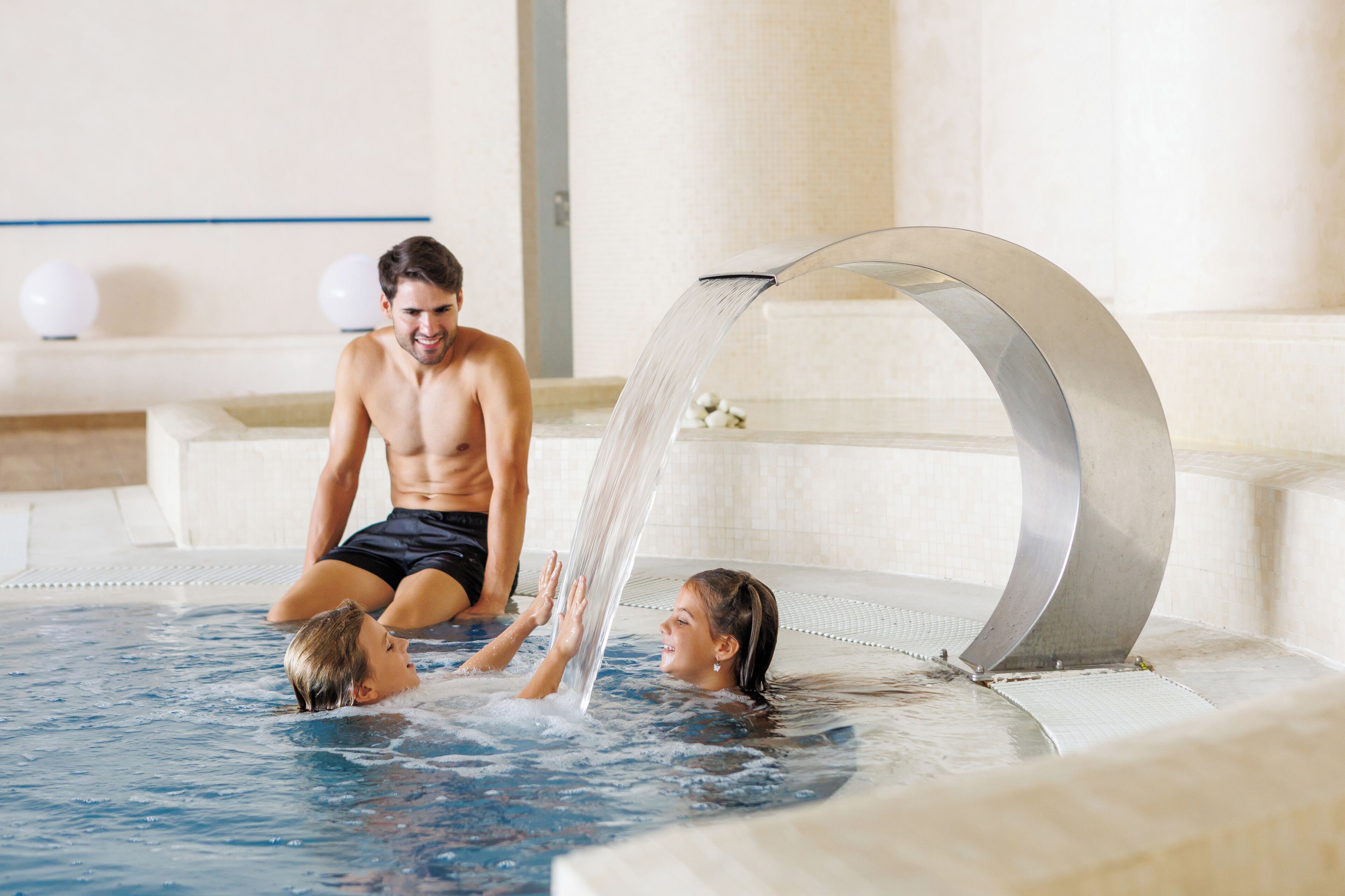 Una piscina con un elemento acuático moderno, una zona de descanso con un muro blanco y una línea azul, y una persona sentada en el borde de la piscina.