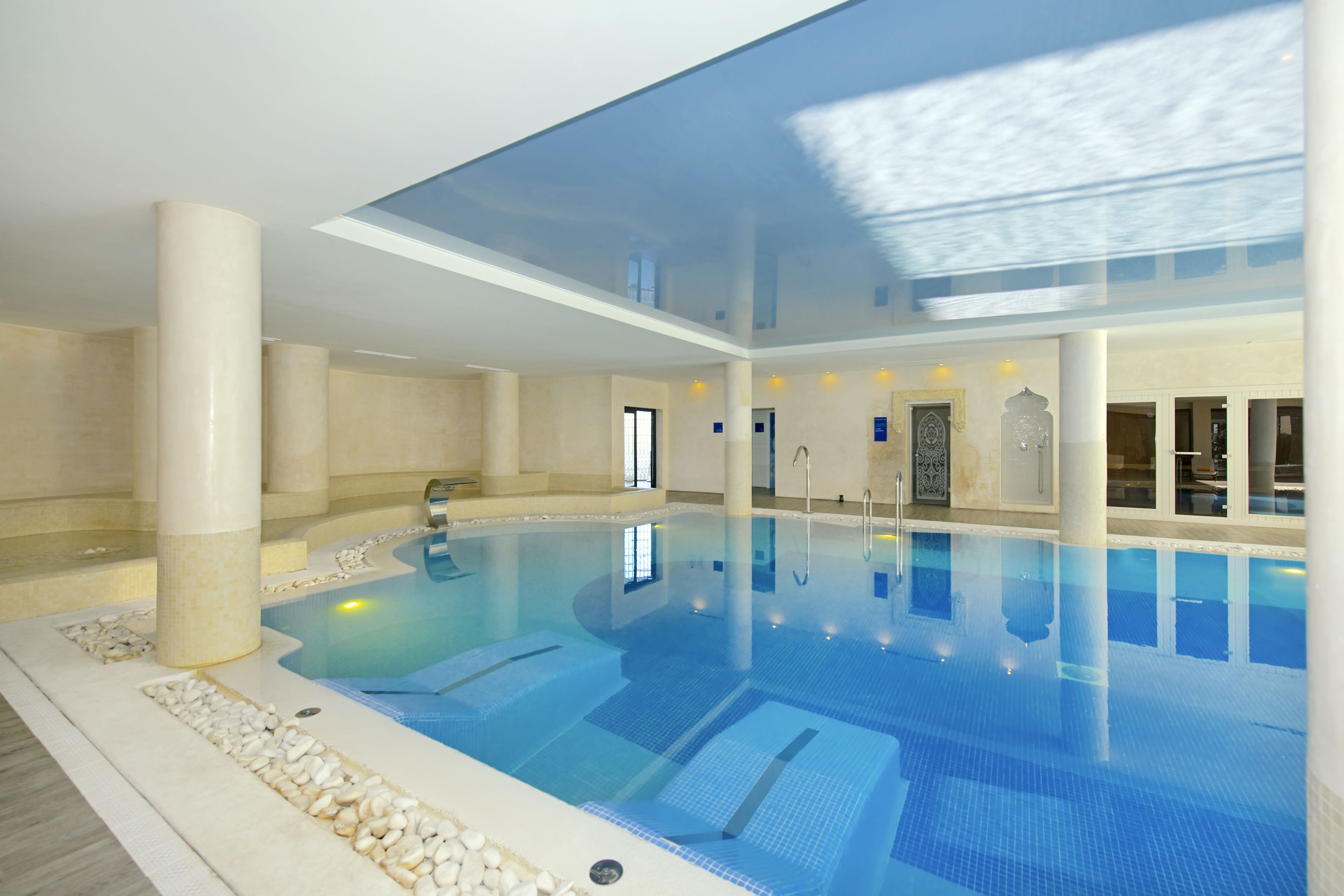 Una piscina interior con agua cristalina, techo de cristal y suelo de baldosas.