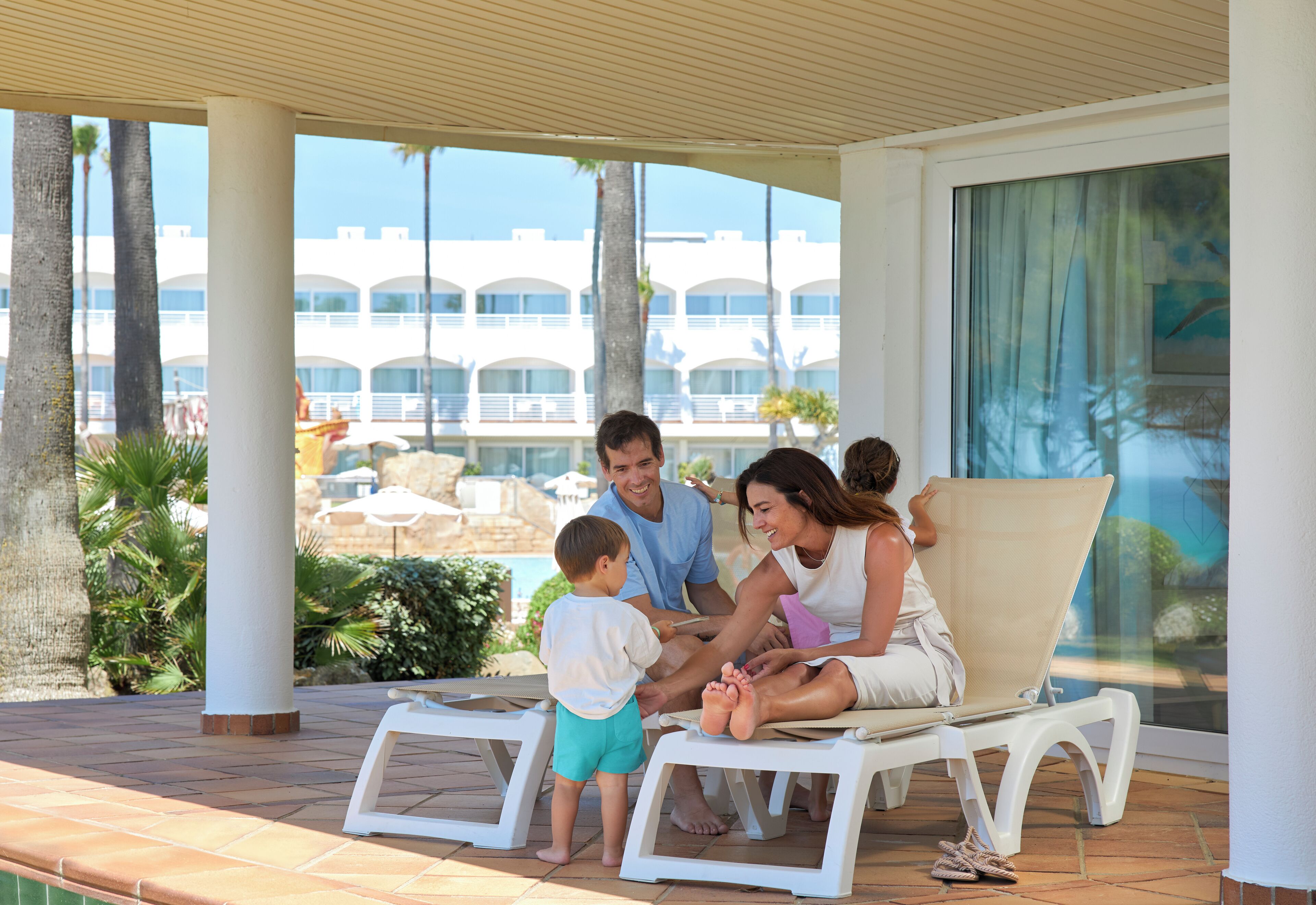 A family on a covered patio with a pool view, white lounge chairs, and a building with a modern design in the background.