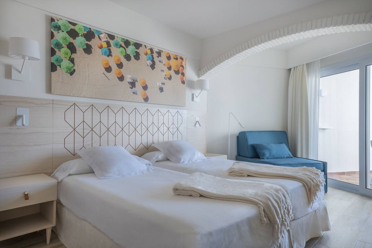 Een moderne hotelkamer met twee bedden, een blauwe bank, een groot abstract schilderij en uitzicht op de omgeving.
