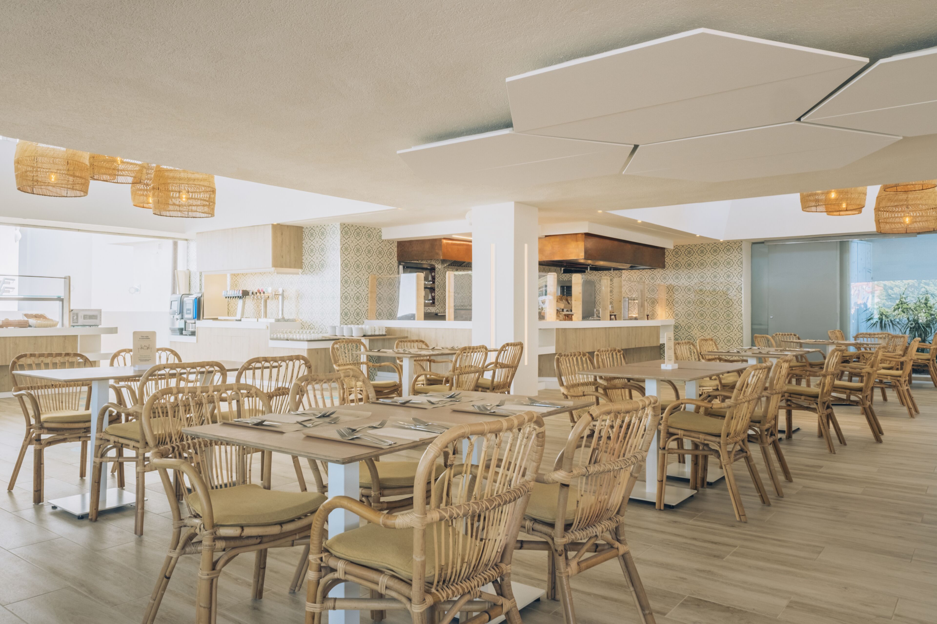 Un comedor amplio con varias mesas y sillas de mimbre, una cocina al fondo y vistas al exterior.